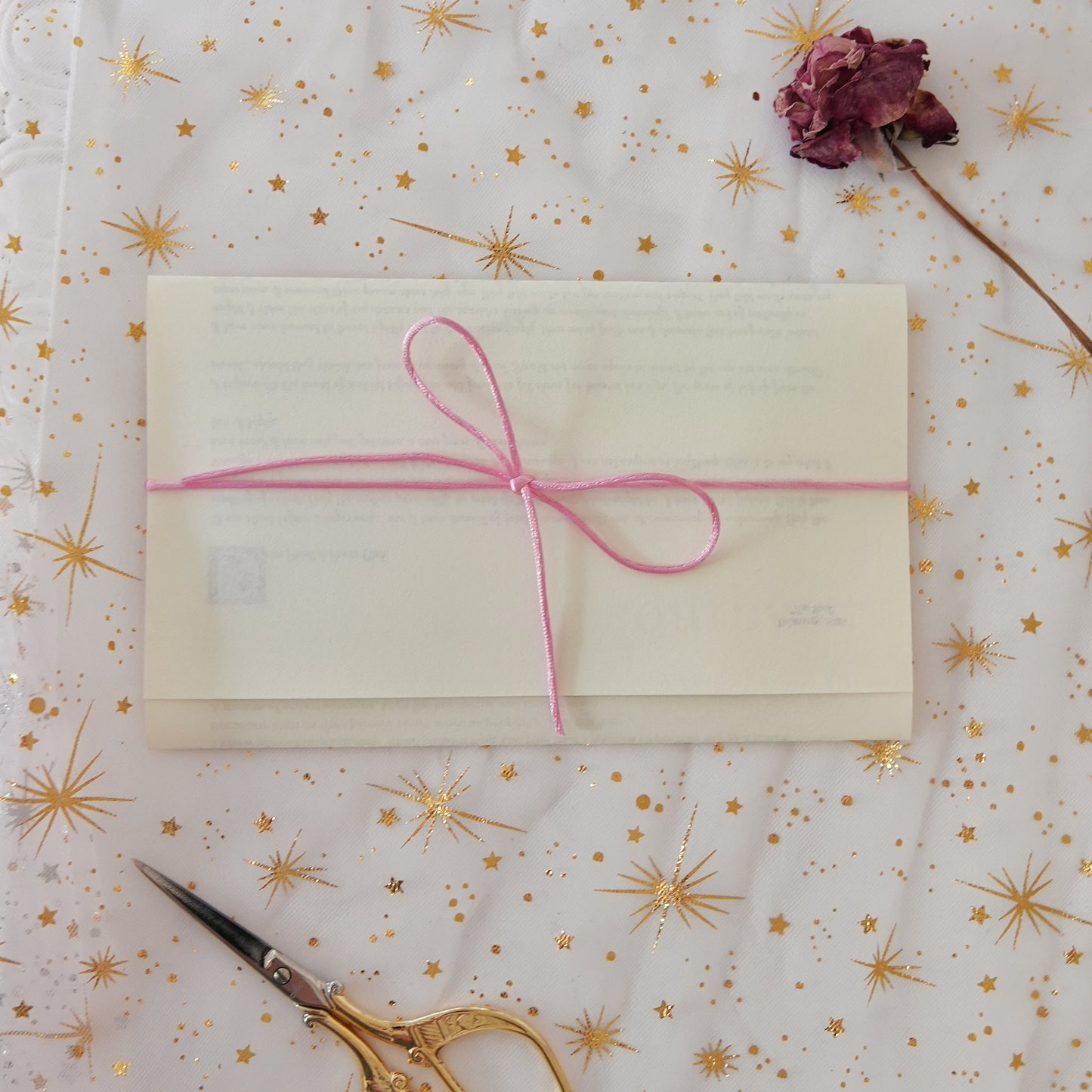 White envelope with pink ribbon on a decorative surface with gold stars and a dried flower.