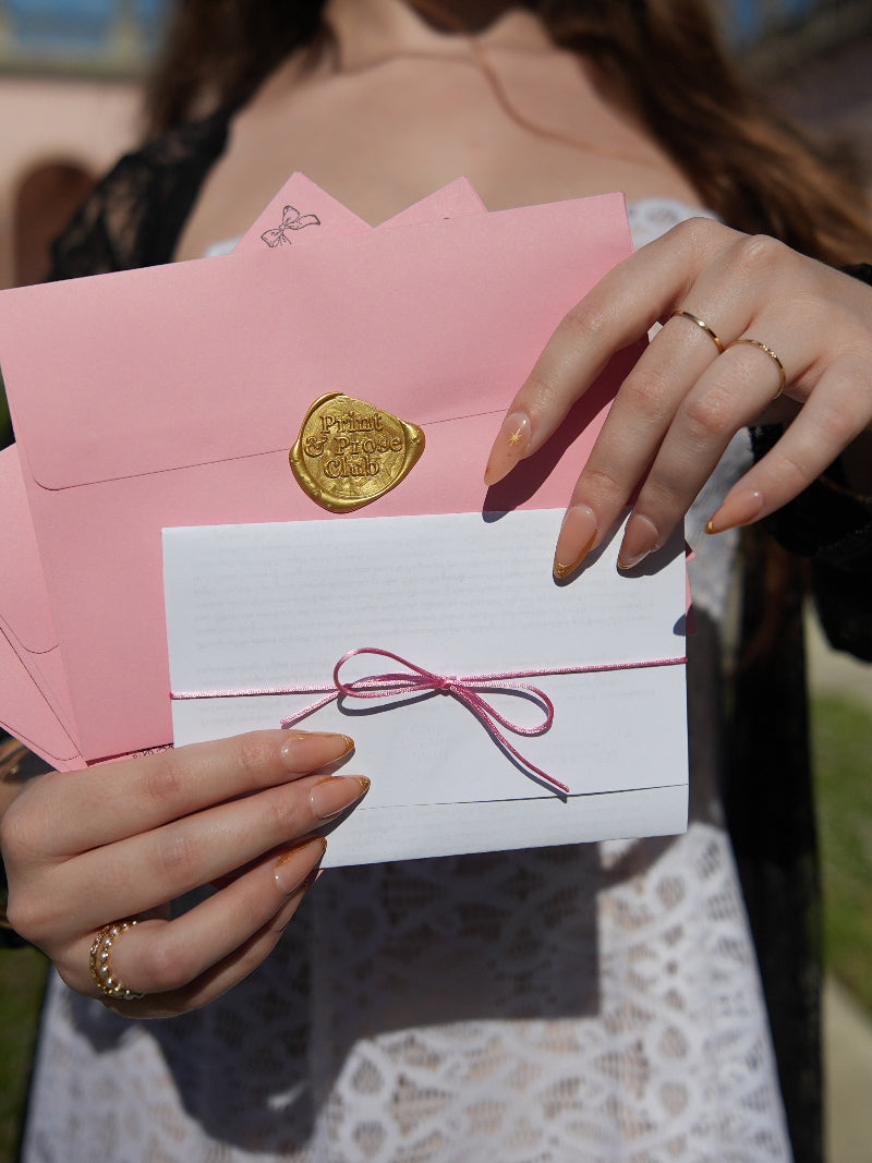 Serenity Blyss holding a pink envelope with a white card featuring a gold seal and pink ribbon for Print & Prose Club.