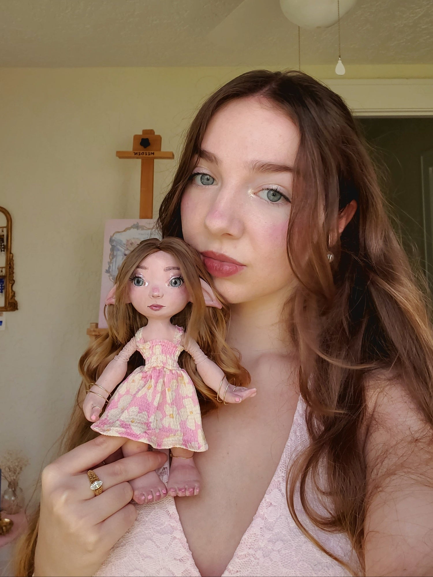 A woman holding a small doll with long brown hair wearing a pink and yellow floral dress, posed in a home interior setting.